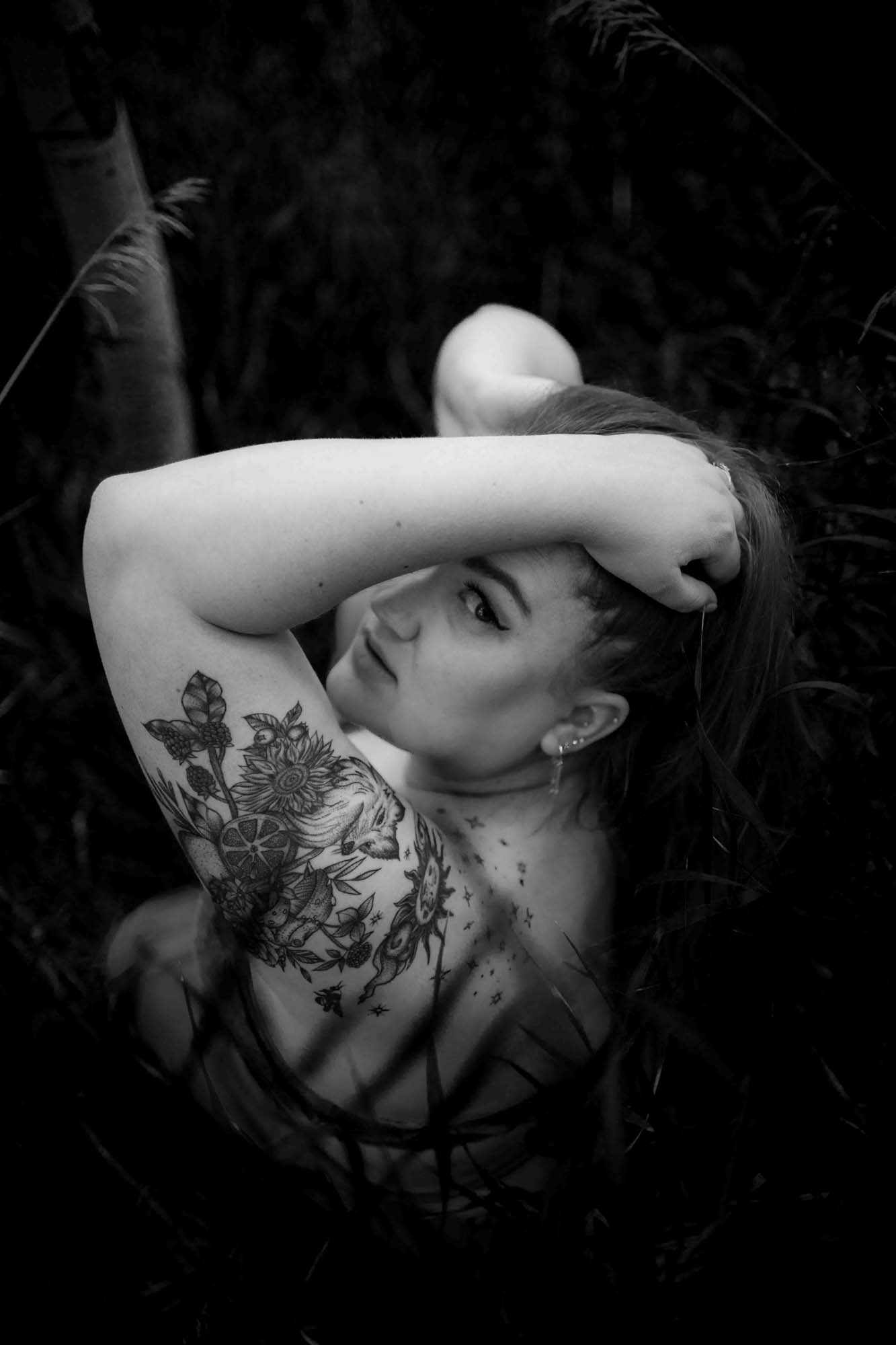 Black and white boudoir portrait of a tattooed woman posing outdoors with her arm raised, captured in a moody, natural setting during a Utah boudoir photography session.