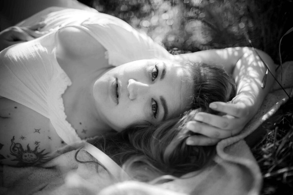 A black and white Utah boudoir shot of a woman lying on the ground, with soft, natural light adding a sultry vibe to the scene. She looks up with a relaxed, sexy expression, her body casually exposed. The shadows play beautifully on her skin, adding to the intimate and irresistible energy of the shot.