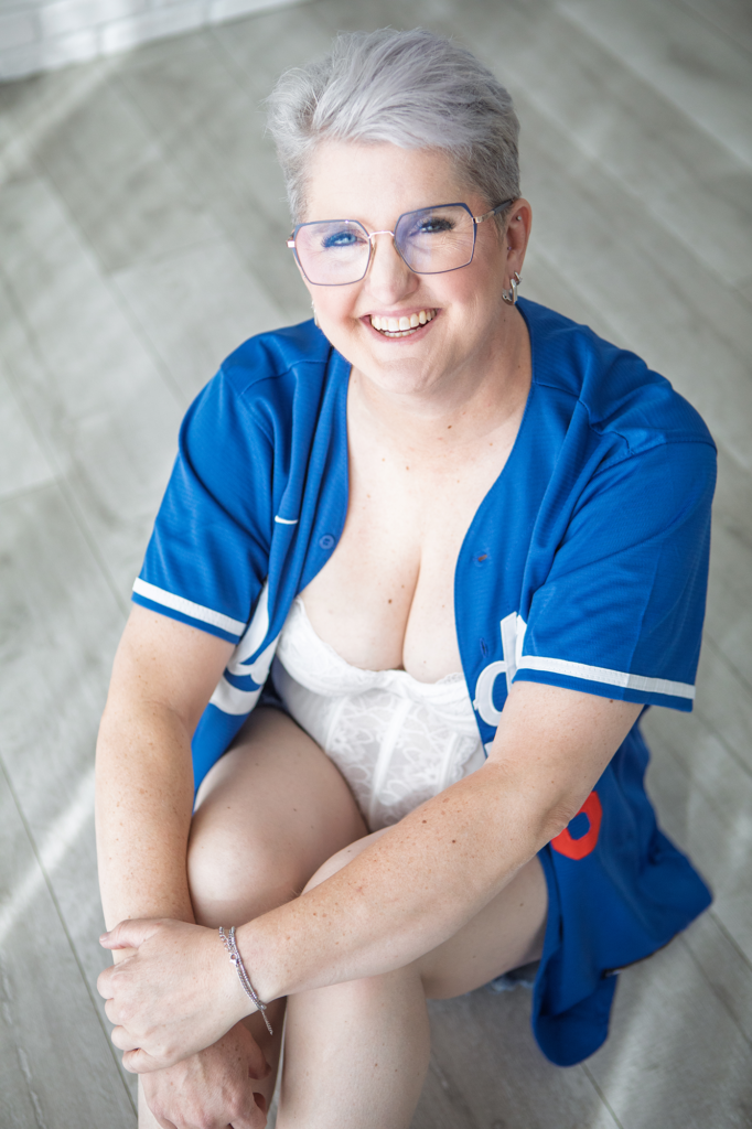 A woman smiling while seated during a boudoir photography session in Utah, wearing lingerie layered with a casual Dodgers jersey.